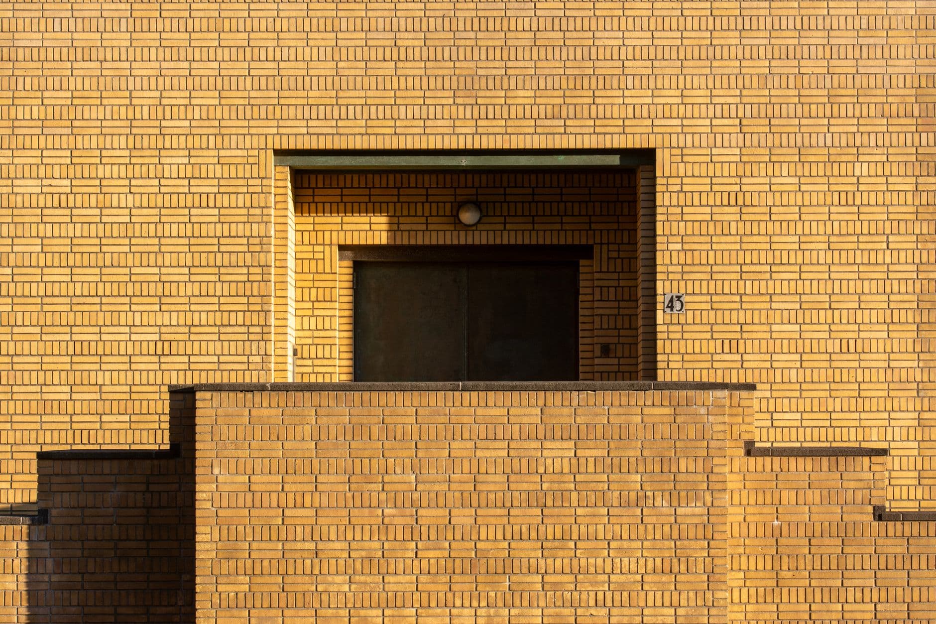 Ochre brick apartment facade in raking sunlight, with a recessed entrance and stepped brick parapet walls.