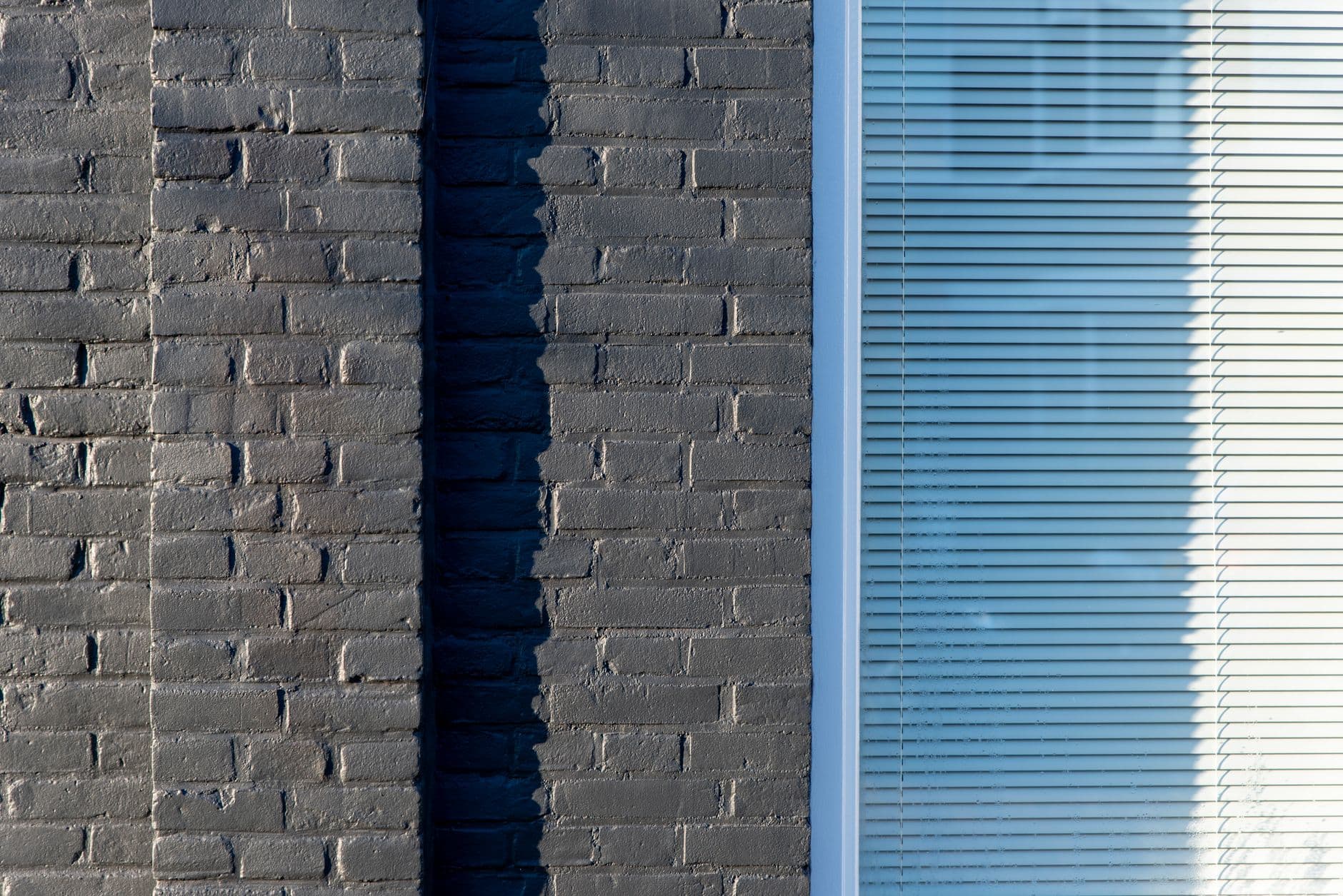 Close view of gray-painted brick pilasters casting a vertical shadow beside a large window with closed horizontal blinds.
