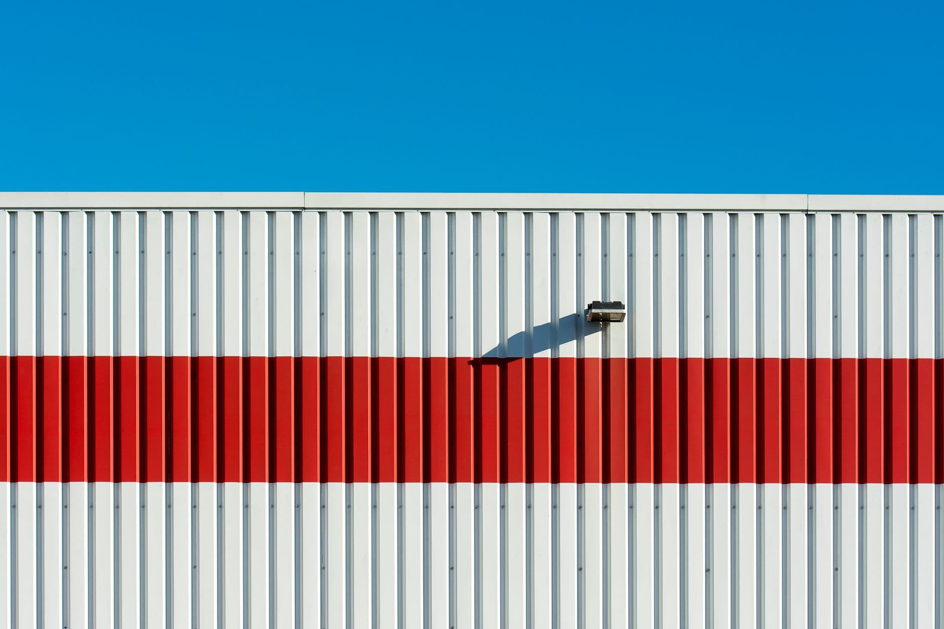 White corrugated warehouse wall crossed by a single horizontal red stripe, with one floodlight casting a diagonal shadow under a clear blue sky.