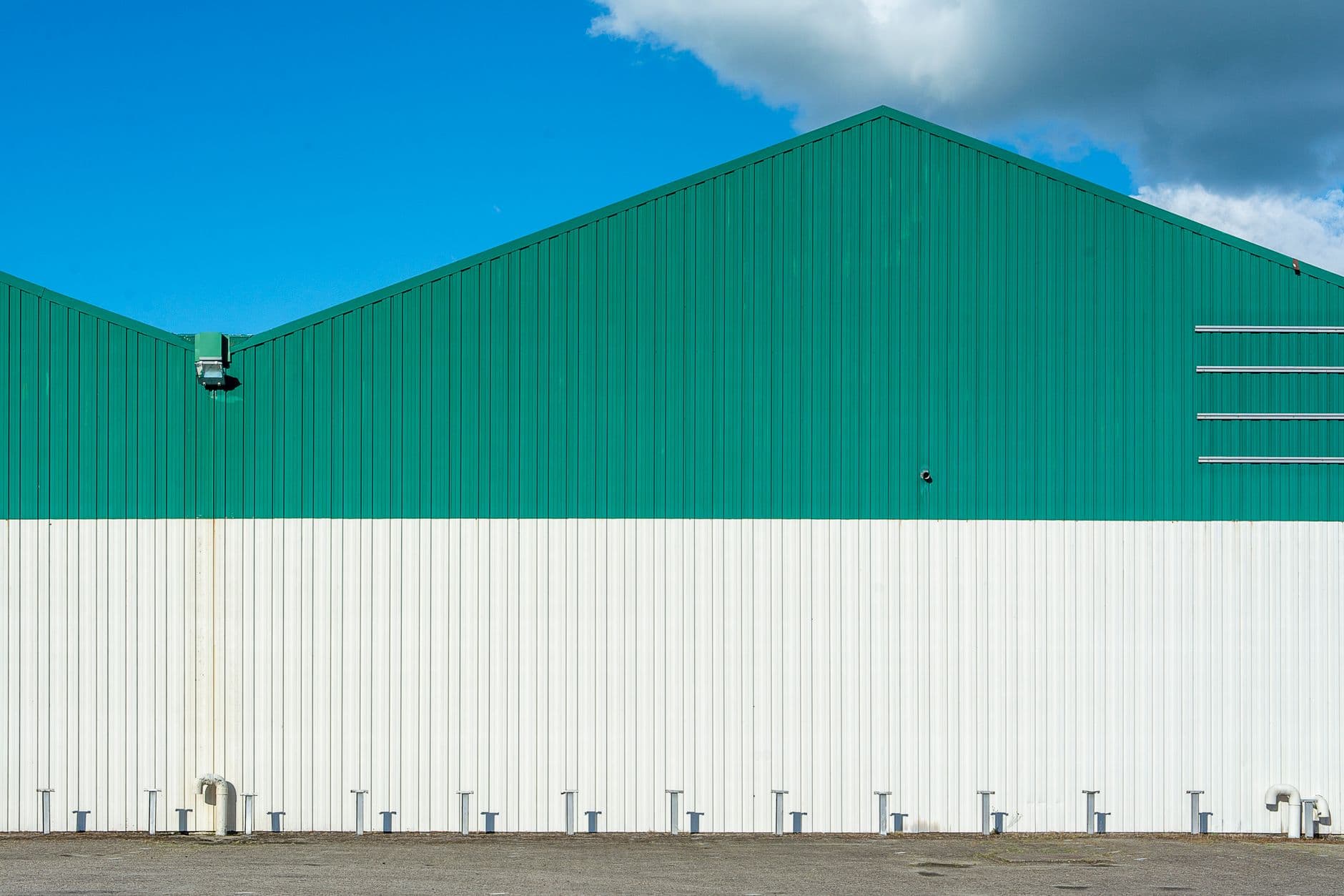Gabled warehouse exterior with green upper cladding over white corrugated walls, photographed frontally against a blue sky.