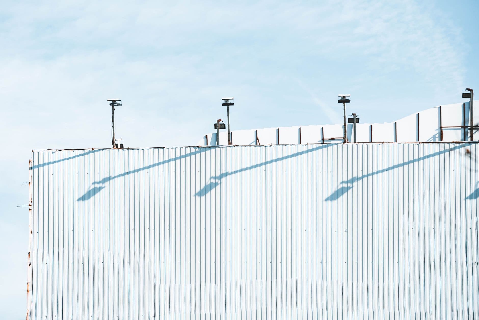 White corrugated industrial wall with a row of roof-mounted floodlights casting long diagonal shadows in bright daylight.